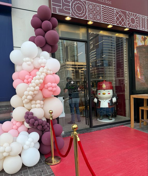 The entrance of an 'EGG' branded store features a large decorative balloon arrangement in purple, pink, and white, with a penguin mascot visible through the glass doors and a red carpet leading inside.