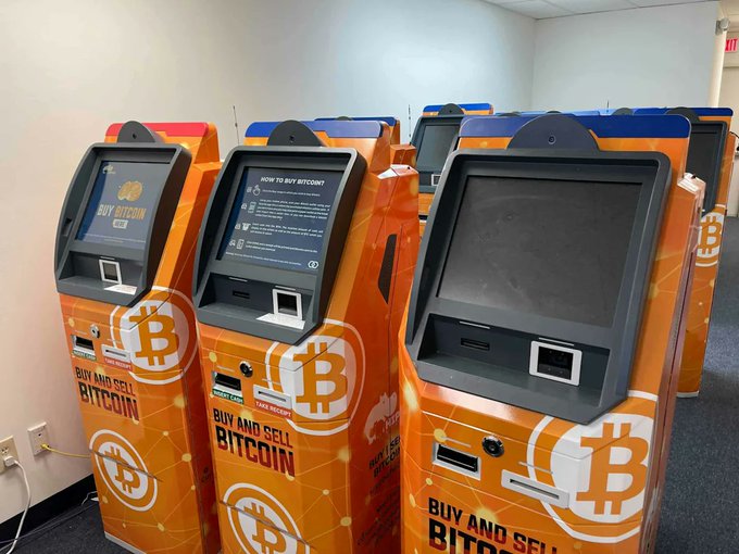 A row of orange Bitcoin ATMs with 'BUY AND SELL BITCOIN' branding and Bitcoin logos, featuring screens displaying 'HOW TO BUY BITCOIN!' instructions.