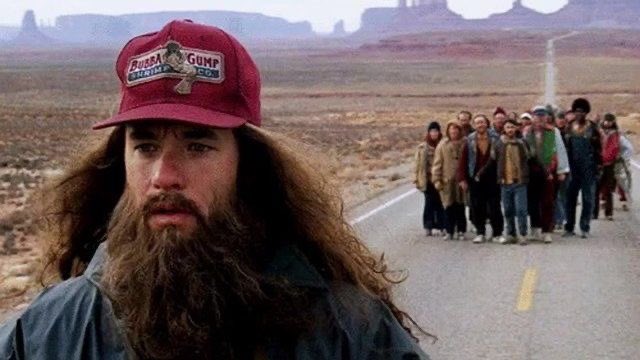 A scene from the movie Forrest Gump, showing the title character in his Bubba Gump cap with his long hair and beard, while a group of people follows him along a desert highway.