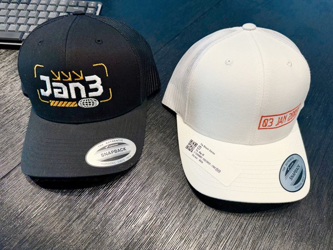 The image features two hats, one black and one white, placed side by side on a wooden surface. The black hat has "JAN3" written on it, while the white hat displays an orange label with the text "03 Jan 2009". Both hats are positioned in front of a computer keyboard, suggesting that they might be for sale or used as promotional items related to a company named "Genes".