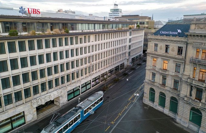An aerial view of a city street showing a UBS bank building on the left and a Credit Suisse building on the right, with a blue tram passing between them.