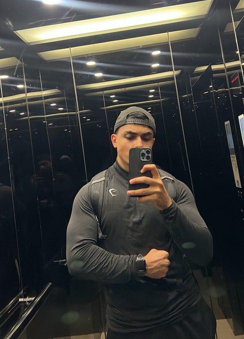 A mirror selfie of a muscular man wearing a gray athletic top and backward cap, flexing his arm while holding a phone in what appears to be a modern elevator with mirrored walls.