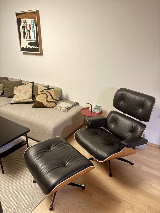 A modern living room featuring a black leather Eames-style lounge chair and ottoman next to a gray sofa with patterned pillows, with framed artwork hanging on the white wall above.