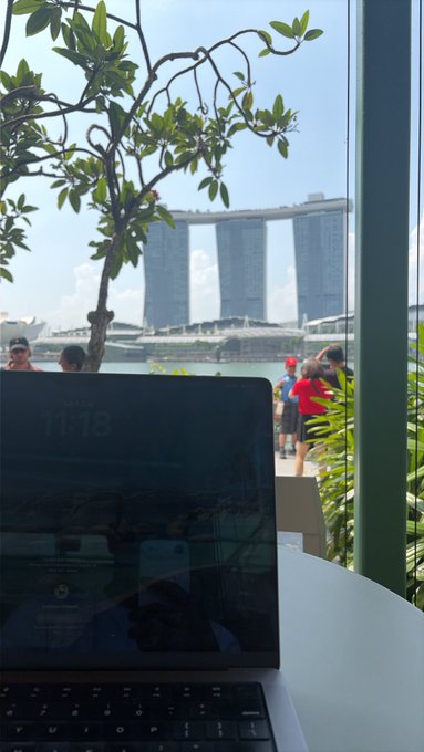 A laptop screen showing the time '11:18' sits on a white table, with a scenic view of the Marina Bay Sands hotel in Singapore visible through a window framed by green foliage.