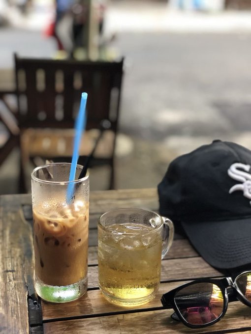 A wooden cafe table with an iced coffee in a tall glass, an iced beverage in a mug, a black Chicago White Sox cap, and sunglasses.