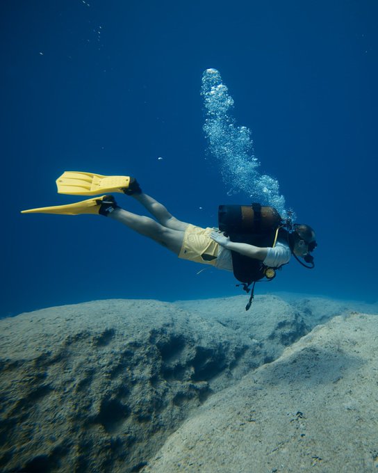 A scuba diver swims over a sandy seabed in clear blue water, wearing yellow fins, yellow swim trunks, and a black scuba tank with bubbles rising above.