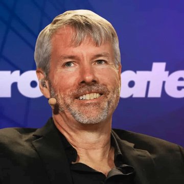 A man with gray hair and a beard speaks into a microphone at an event with a blue background. The image accompanies news about Michael Saylor's bullish prediction that Bitcoin ($BTC) will reclaim the $100,000 level.