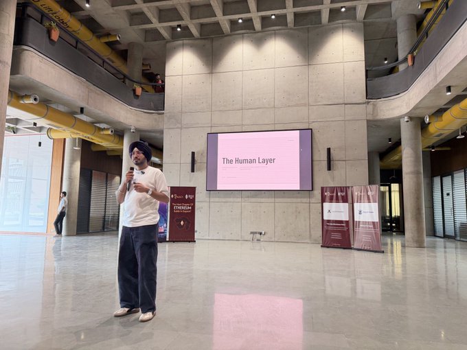 A man with a turban holds a microphone in a modern, spacious hall. Behind him, a screen displays the text 'The Human Layer' in pink.