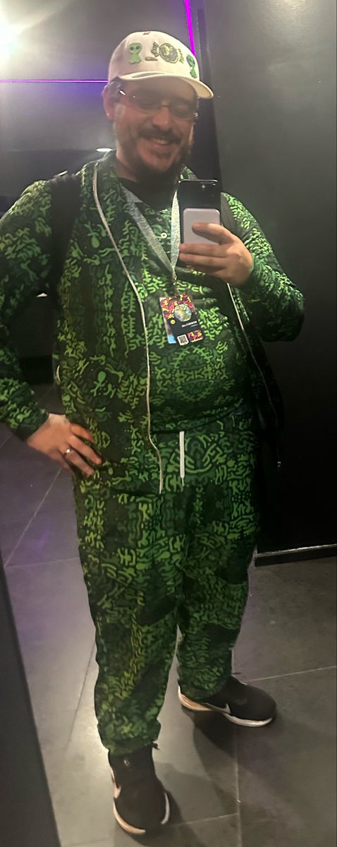 A man takes a mirror selfie wearing a green patterned matching set, white cap with pins, glasses, and a backpack strap, while holding his smartphone.