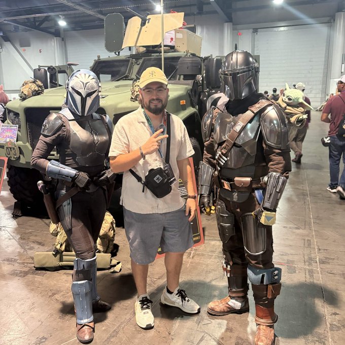 A man posing with two people in Mandalorian armor costumes at what appears to be a comic convention, with a military vehicle visible in the background.