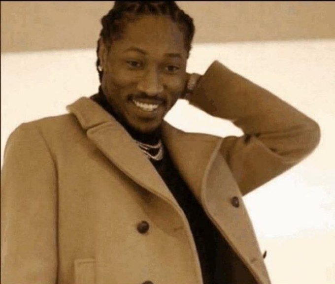 A smiling man with braided hair wearing a tan coat and black turtleneck, posing with his hand behind his head.