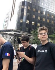 Two young men standing outdoors in front of a tall building with lit windows; one wears glasses and an "EVOLVE" t-shirt, while the other in front is looking down at a phone.