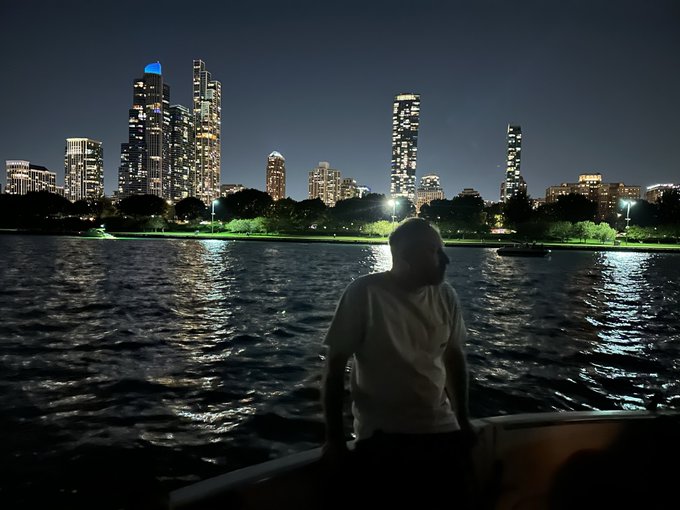 A nighttime silhouette of a person on a boat with a city skyline of illuminated skyscrapers across the water.