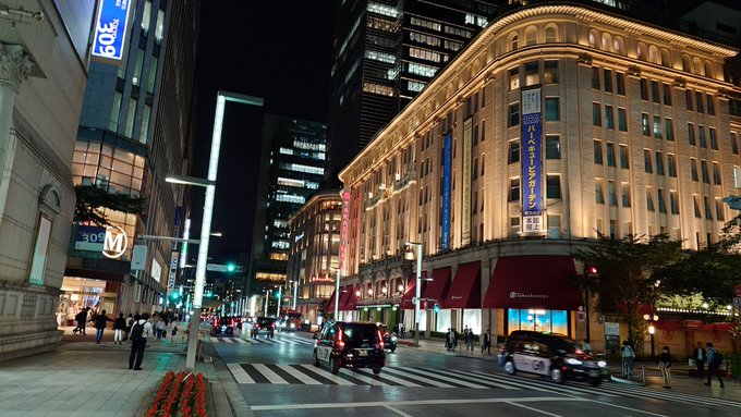 The image shows a busy city street at night with tall buildings lining both sides, including a large building that has a blue sign on its side. The street is filled with cars driving down it, and there are several people walking along the sidewalks. A traffic light can be seen in the foreground of the photo.