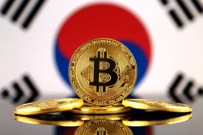 A Bitcoin token in the foreground with the South Korean flag blurred in the background, alongside several gold coins on a reflective surface.