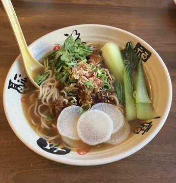 A bowl of Asian noodle soup is garnished with sliced radishes, greens, cilantro, and a savory meat topping, served with a spoon.