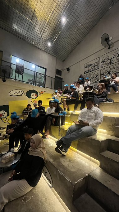 A group of people are gathered on tiered seating, some wearing blue hats, while a cartoon mural is visible on the wall, hinting at a lively gathering related to "Ethereum Genesis: Quantum Leap."