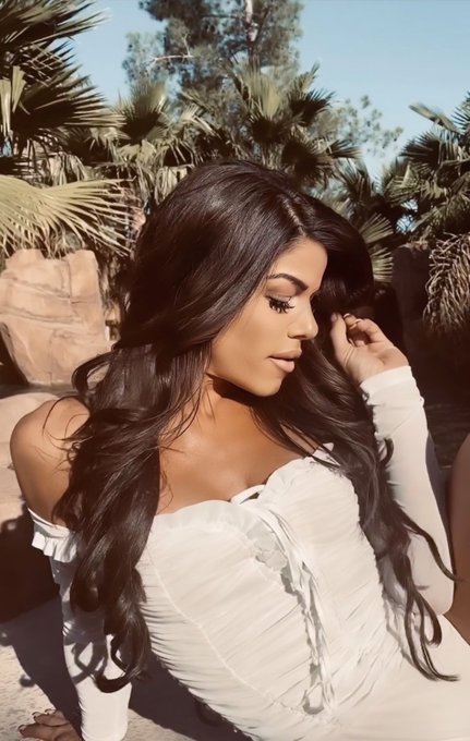 A woman with long, wavy dark hair wearing a white off-the-shoulder top poses in an outdoor setting with palm trees in the background.