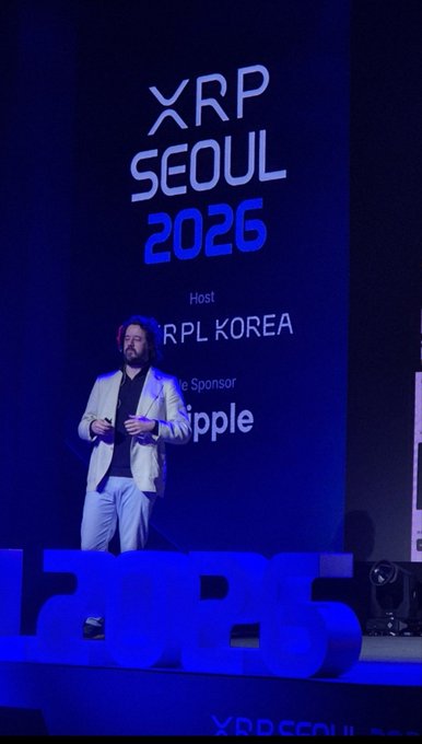A presenter speaks on stage at the XRP Seoul 2026 conference, with the event's logo displayed prominently behind him alongside sponsor branding for Ripple, as the image illustrates the real-world XRP ecosystem utility being highlighted in the accompanying post about Tria's self-custody financial services.