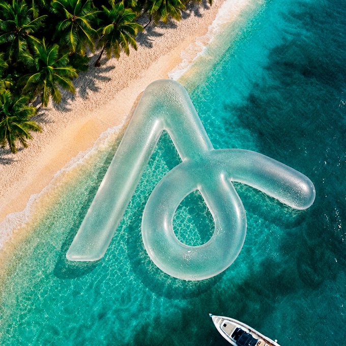 An aerial view of a tropical beach with turquoise water and palm trees, featuring a giant translucent inflatable 'A' letter logo floating in the shallow water near a small boat.