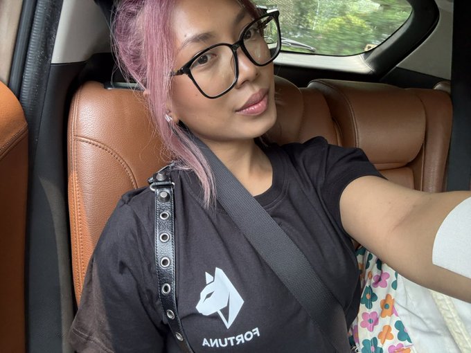 A selfie of a young woman with pink hair and black glasses sitting in the passenger seat of a car with tan leather interior, wearing a black Fortuna t-shirt.