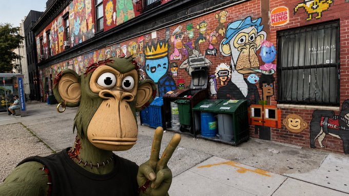A person in a Bored Ape NFT costume takes a selfie while flashing a peace sign in front of the famous NFT wall in New York City, which is covered in colorful murals of ape characters and crypto art including "THE KING" signage, with recycling bins lining the sidewalk in front of the brick building.