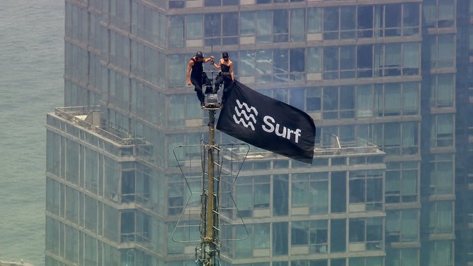 A person climbs a tower to plant a black flag emblazoned with the "Surf" logo, set against a backdrop of a towering glass building—a fitting visual metaphor for the poster's claim of being an early-morning "surfer" of the markets, scanning overnight news to stay above the noise.