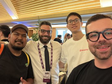 A selfie of four smiling men at what appears to be a conference or event, with two wearing lanyards and one wearing a 'Devcon' branded white shirt.