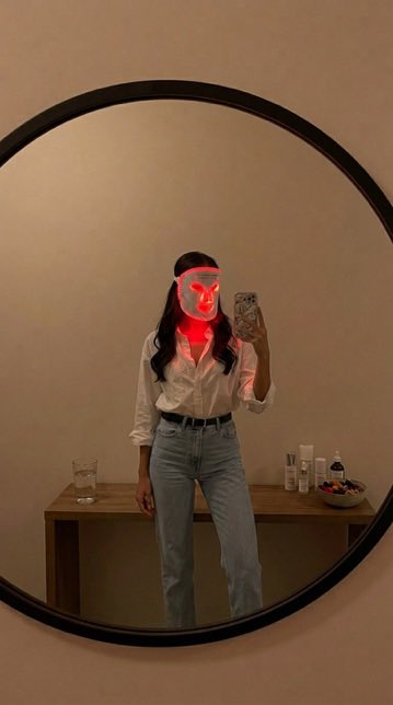 A mirror selfie of a woman wearing a white shirt and light blue jeans, with a red LED light therapy mask on her face.