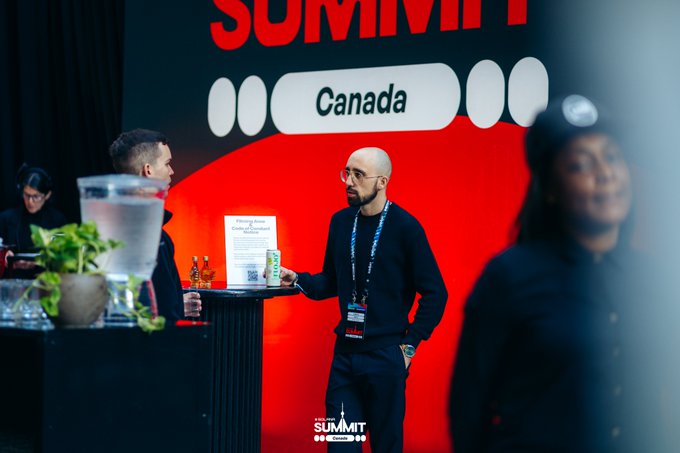 Two men converse at the 'Ads Con Canada Summit' booth, with one holding a beverage can and promotional bottles displayed on the table.
