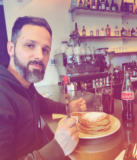 A bearded man in a brown hoodie sits at a cafe counter holding a knife and fork, ready to eat a stack of pancakes next to a glass Coca-Cola bottle.