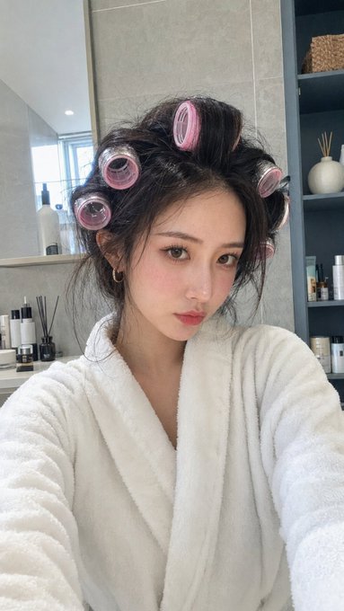The image features a young woman wearing a white robe, with her hair styled into curls using pink curlers. She appears to be posing for a photo or selfie, as indicated by her expression. The background of the image includes various bottles scattered around, suggesting that she might have been in a bathroom or dressing area where these items are commonly found.