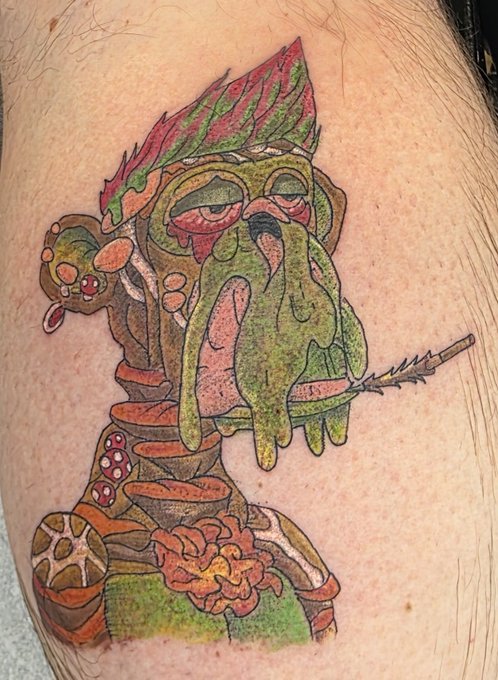 A colorful cartoon-style tattoo on skin depicting a zombie-like ape with green dripping skin, spiked red hair, and X-ray vision eyes holding a feather, fitting the "BAYC FOR LIFE" Bored Ape Yacht Club reference. The chaotic, drippy aesthetic directly mirrors the distinctive art style of the popular BAYC NFT collection.