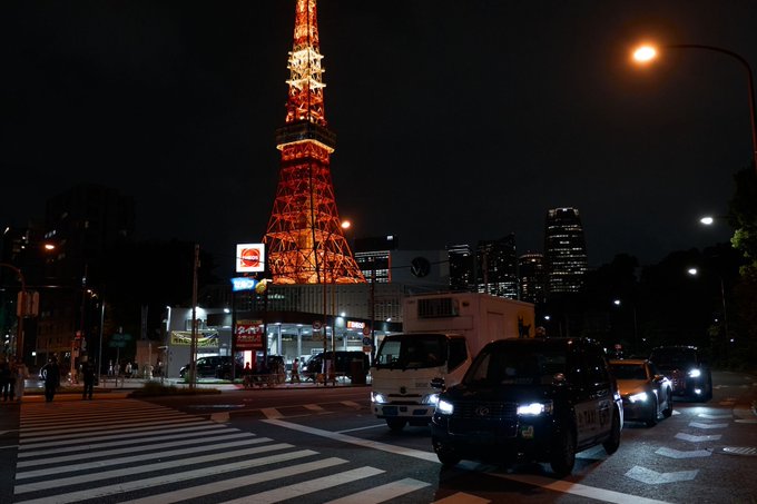 A nighttime cityscape featuring the illuminated orange Tokyo Tower, with vehicles including taxis and a white truck stopped at a crosswalk in the foreground.