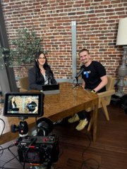 A behind-the-scenes photograph of a video podcast setup, showing a man and a woman seated at a wooden table with laptops and a microphone, with a camera on a tripod filming them in a brick-walled studio.