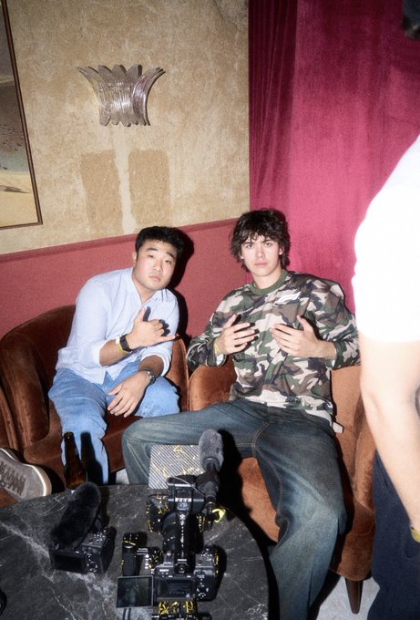 Two young men pose for a photo while seated on a brown couch, one in a light blue shirt and the other in a camouflage top, with camera equipment visible in the foreground. The casual behind-the-scenes shot captures a relaxed, candid moment that reflects the "GOAT" (Greatest of All Time) sentiment, highlighting the camaraderie and confidence of the subjects being celebrated.