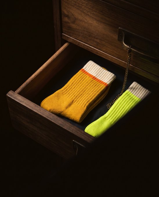 A dark wooden drawer is partially open, revealing a folded pair of yellow-orange socks with white cuffs and a single neon green sock with a white cuff inside, tied with a chain that appears to be a key or lock fob—visually suggesting "cross-chain" connectivity, as if the socks themselves were linked or chained across different chains, mirroring the post's announcement about Cross Chain Tokens (CCT) launching on Sock across BSC and Robinhood Chain.