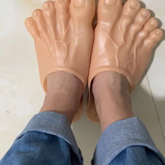 A person wearing jeans and novelty slippers shaped like bare human feet stands on a tiled floor.