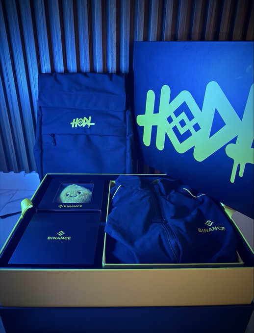 A Binance promotional package containing a backpack, tracksuit, notebook, and small plush figure, with a yellow 'HOON' logo graffiti-style design on the box.