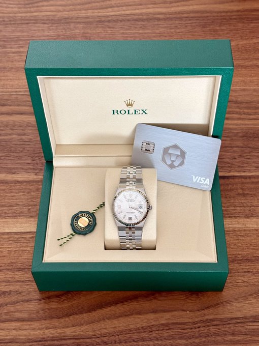 An open green Rolex presentation box displaying a stainless steel Rolex Datejust watch on a cream cushion, accompanied by a metal Visa credit card with a logo and a green Rolex hangtag.