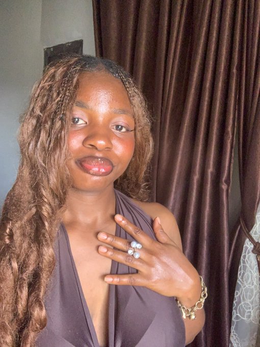 A woman with long braided hair poses indoors wearing a gray top and rings.