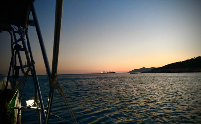 A serene sunset view from a sailboat shows calm waters, a distant cruise ship on the horizon, and the silhouette of a coastline, capturing the tranquil end of a day's journey at sea. The peaceful maritime scene complements the optimistic trading message of looking forward to tomorrow's opportunities.