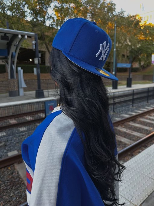A person with long black hair wearing a blue NY Yankees cap and a blue and white jacket, standing at a railway station platform with trees and tracks in the background.