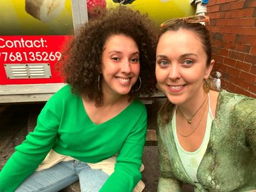 Two women smiling for a selfie, one with curly hair wearing a green shirt and the other with straight hair wearing a green cardigan, in front of a yellow truck with "768135269" visible.