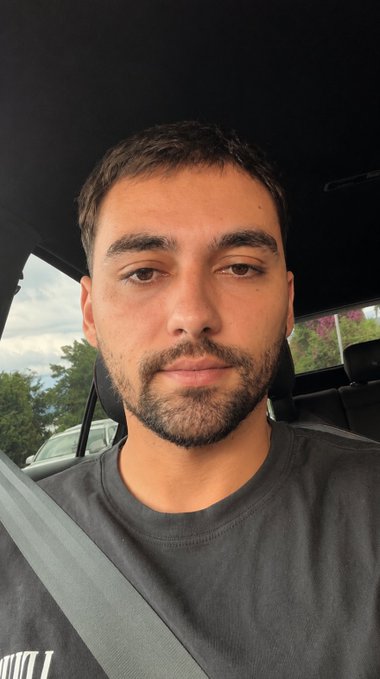 A man with a beard and short hair wearing a dark t-shirt and a seatbelt is shown in this selfie taken in a car. The caption "Can I get a GM?" is a common way to greet someone in the morning, and the image of the man likely represents the person posting the greeting.