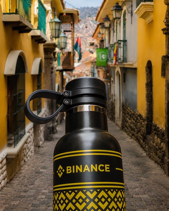 A black "BINANCE" branded water bottle sits on a cobblestone street in what appears to be La Paz, Bolivia, with colorful colonial-style buildings and yellow lamp posts lining the narrow alley. The post asks who recognizes this street, suggesting the image is a visual riddle inviting viewers to identify the iconic Bolivian location.