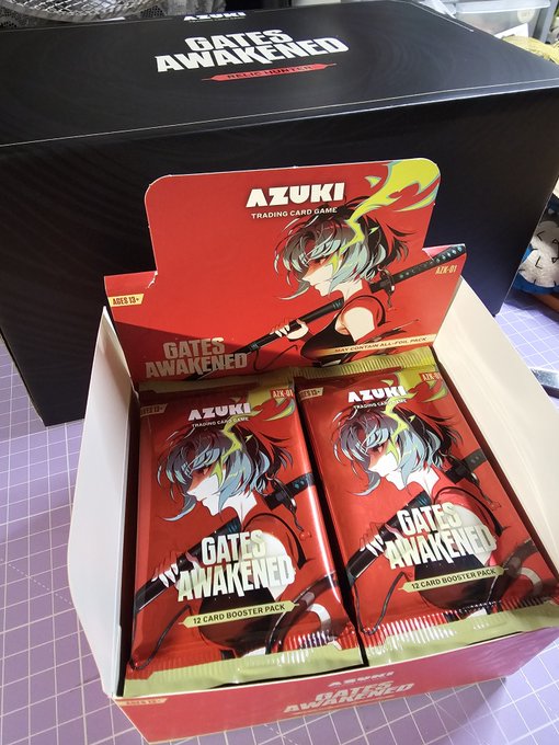 A display box of AZUKI "GATES AWAKENED" trading card game booster packs, containing individually wrapped 12-card booster packs featuring anime character artwork.