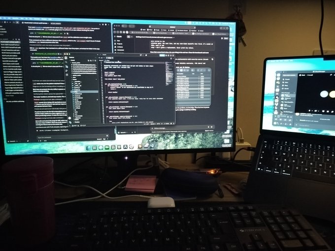 Dimly lit desk setup featuring a large monitor and laptop displaying code windows, with a keyboard and mug in the foreground.