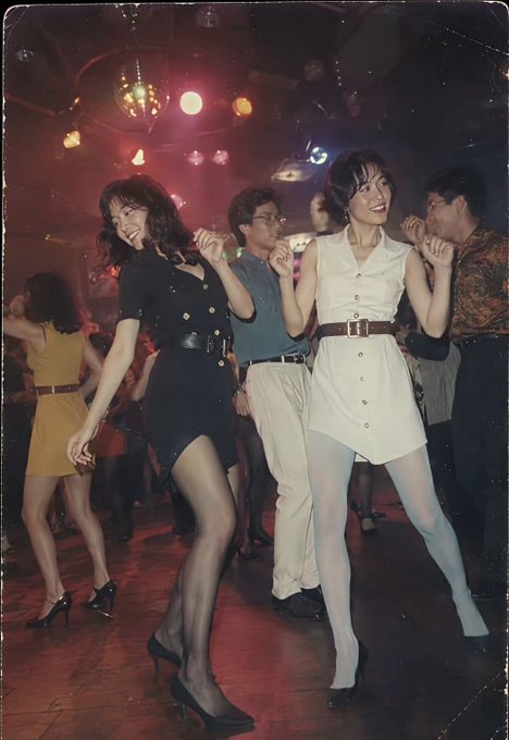 A vintage photo of people dancing at a club with disco lights, featuring two smiling women in the foreground wearing a black dress and a white dress with belts.