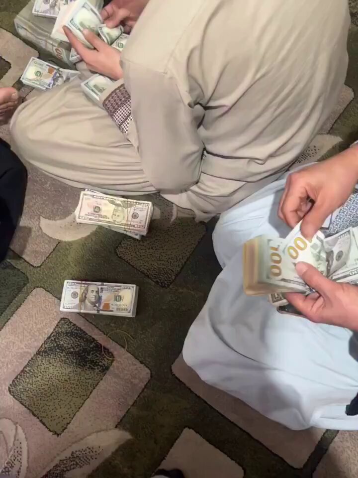 The image shows a group of people sitting on the floor, each holding stacks of US dollar bills. They are all wearing beige clothing, which suggests that they might be participating in an event or gathering related to money or finance. The presence of these stacks of money indicates that it could be a fundraiser or a financial-related activity taking place among the group members.